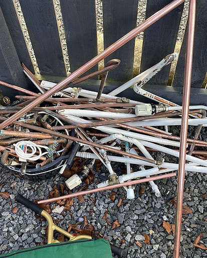 A pile of copper and white metal pipes, with valves and fittings, lies on gravel near a wooden fence.