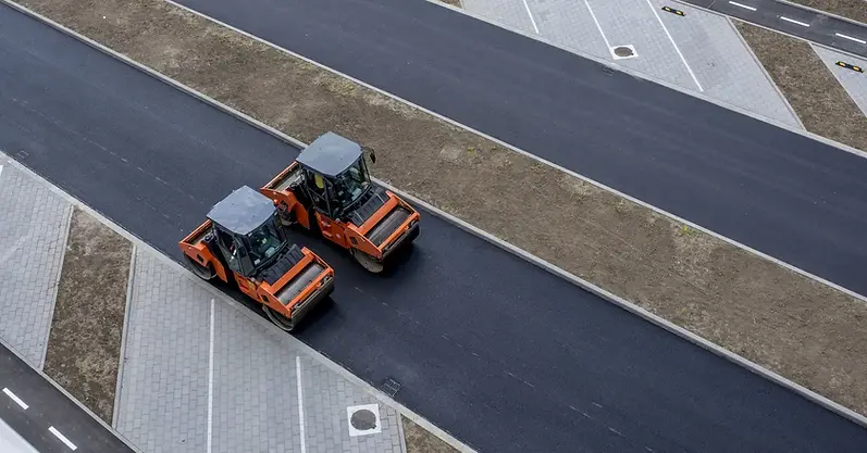 Road roller vehicle compacting a wide stretch of new grey tarmac surface