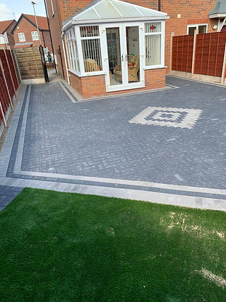 A brick patio in front of a house, bordered by a lush grassy area, creating a welcoming outdoor space
