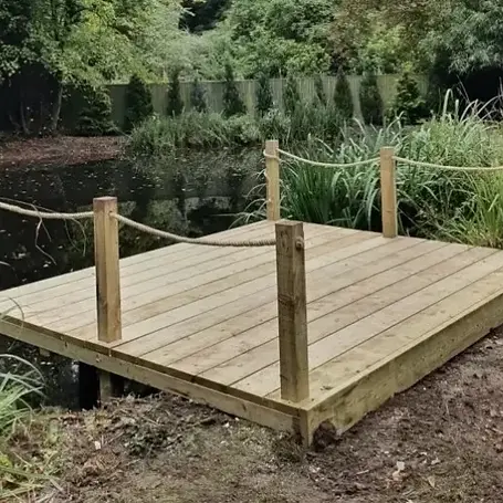 A wooden platform with rope railing stands on the edge of a tranquil pond