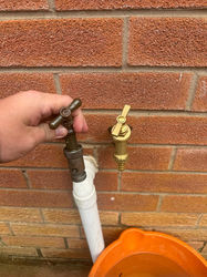 A hand turns an outdoor tap on a brick wall, with a newly installed brass tap beside it