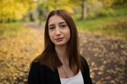 Woman with long brown hair in a black blazer and white top in a blurred park with golden autumn foliage