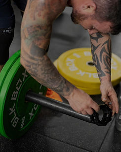 A muscular man with tattoos adjusts a clip on a barbell loaded with green and yellow weights at a gym