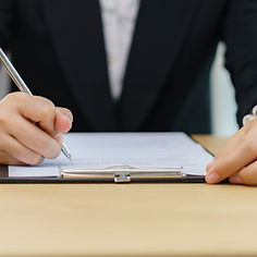 A person in a black suit writes on a clipboard at a light wooden desk.