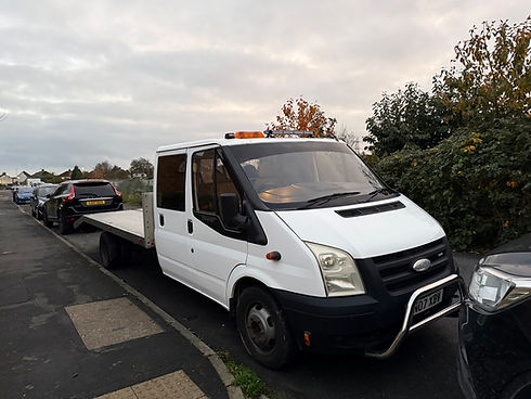 A white flatbed tow truck is parked on a residential street beside a leafy hedge