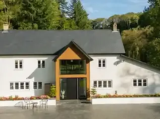 Modern white house with an orange wood gabled entrance and grey roof nestled in dense green woodland