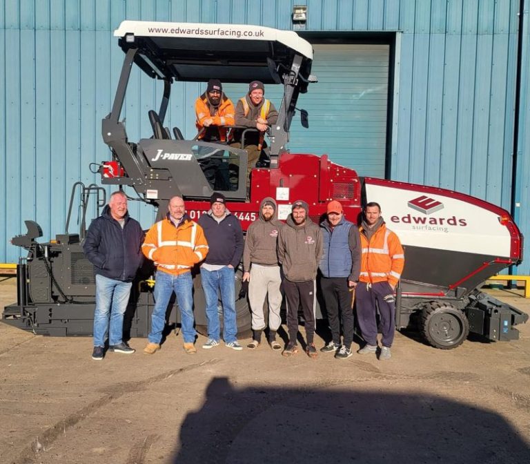 A group of men stands beside a red and white machine, engaged in conversation and examining its features