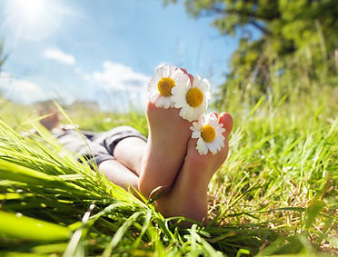 Bare feet rest on lush grass, adorned with daisies between toes under a bright, sunny sky