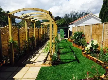Wooden garden pergola over a paved pathway with lush green lawn and flowerbeds under a cloudy sky