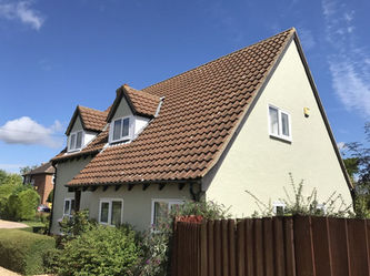 A charming house with a steep red-tiled roof and cream walls, surrounded by greenery and a wooden fence