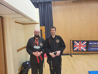 Two men in martial arts uniforms stand smiling in a room with wooden floors and a Union Jack flag
