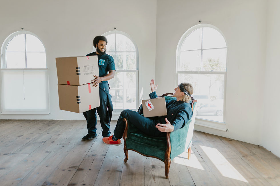 2 men holding moving boxes in an empty living room with one couch. one man is sitting comfortably on the couch.