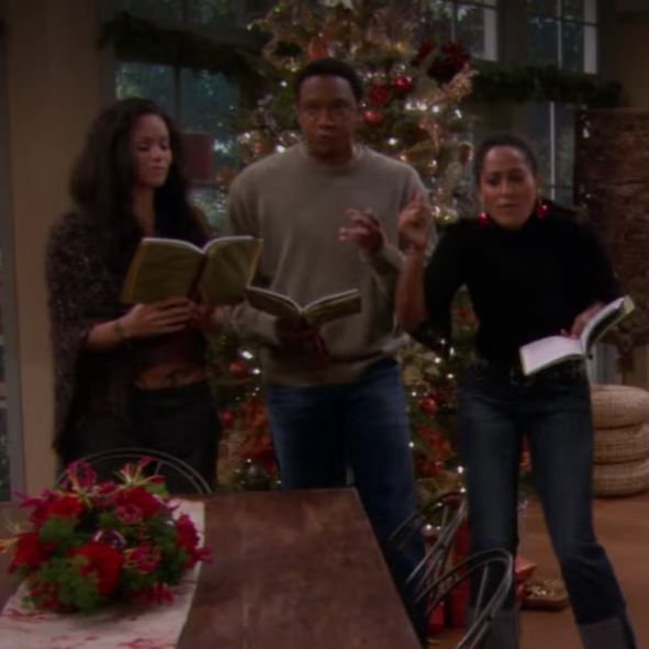 A photo of the TV show "Girlfriends" characters, from left to right: William Dent, Joan Clayton, and Maya Wilkes. They are all wearing festive holiday attire and smiling together in a living room decorated with Christmas decorations, including a Christmas tree in the background.