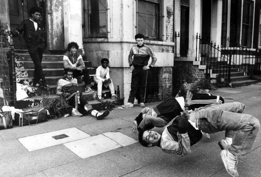 A group of young men, dressed in tracksuits, gather in front of an urban building while one of them performs a backflip on the sidewalk, showcasing their breakdancing skills.