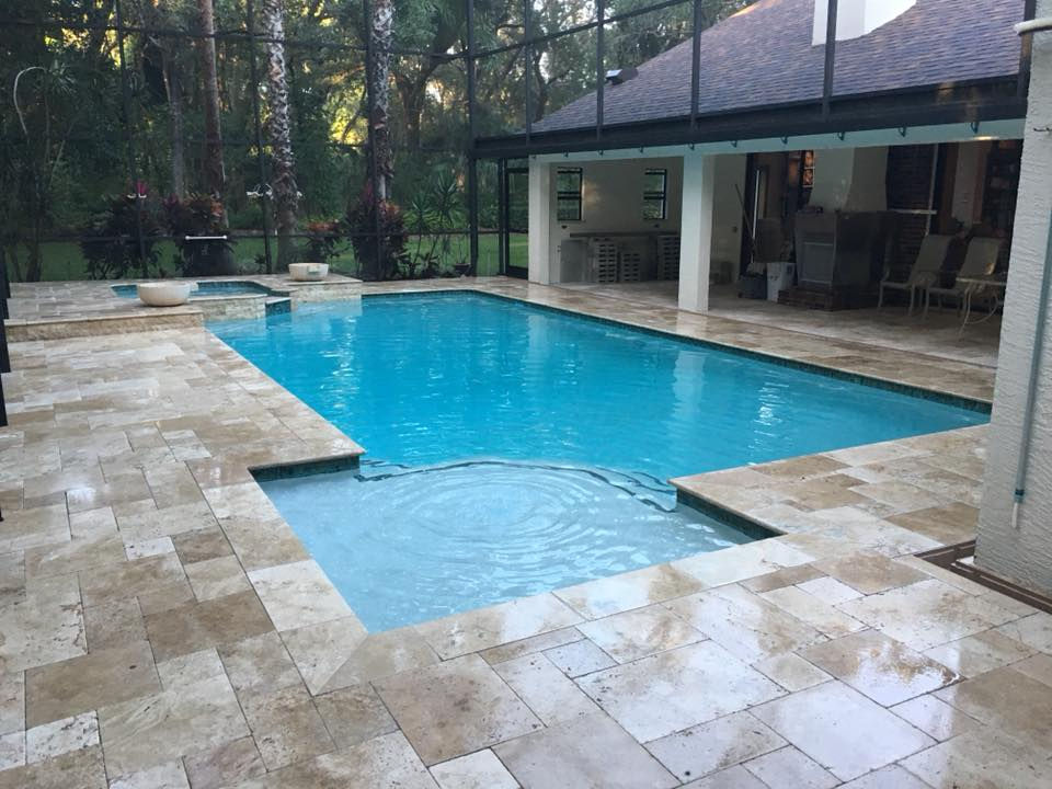 Screen-enclosed rectangular pool with a broad entry shelf, raised spa and light stone decking beside a covered patio.