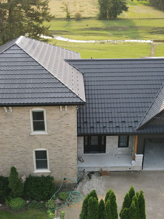 Aerial view of a renovated farmhouse with a dark Mastershake metal roof, blending classic brickwork and modern siding, set against a scenic rural landscape.
