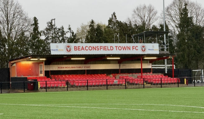 Red stadium seats with "Beaconsfield Town FC" sign on top. Green field in foreground, trees in background, overcast sky.
