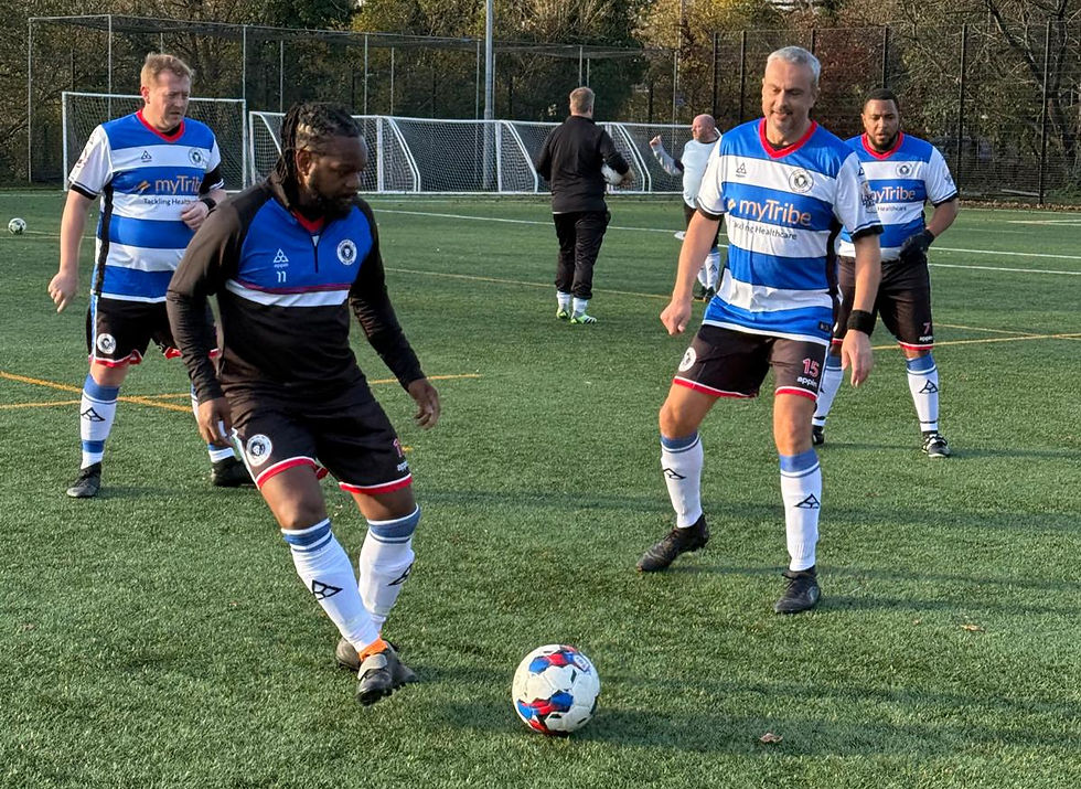 Football players in blue and white jerseys on a grassy field. One dribbles the ball while others observe. A net and trees in the background.