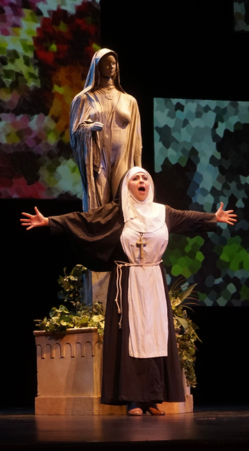 Victoria sings onstage in a nun habit with her arms out to her sides. There is a female nun statue in the background.