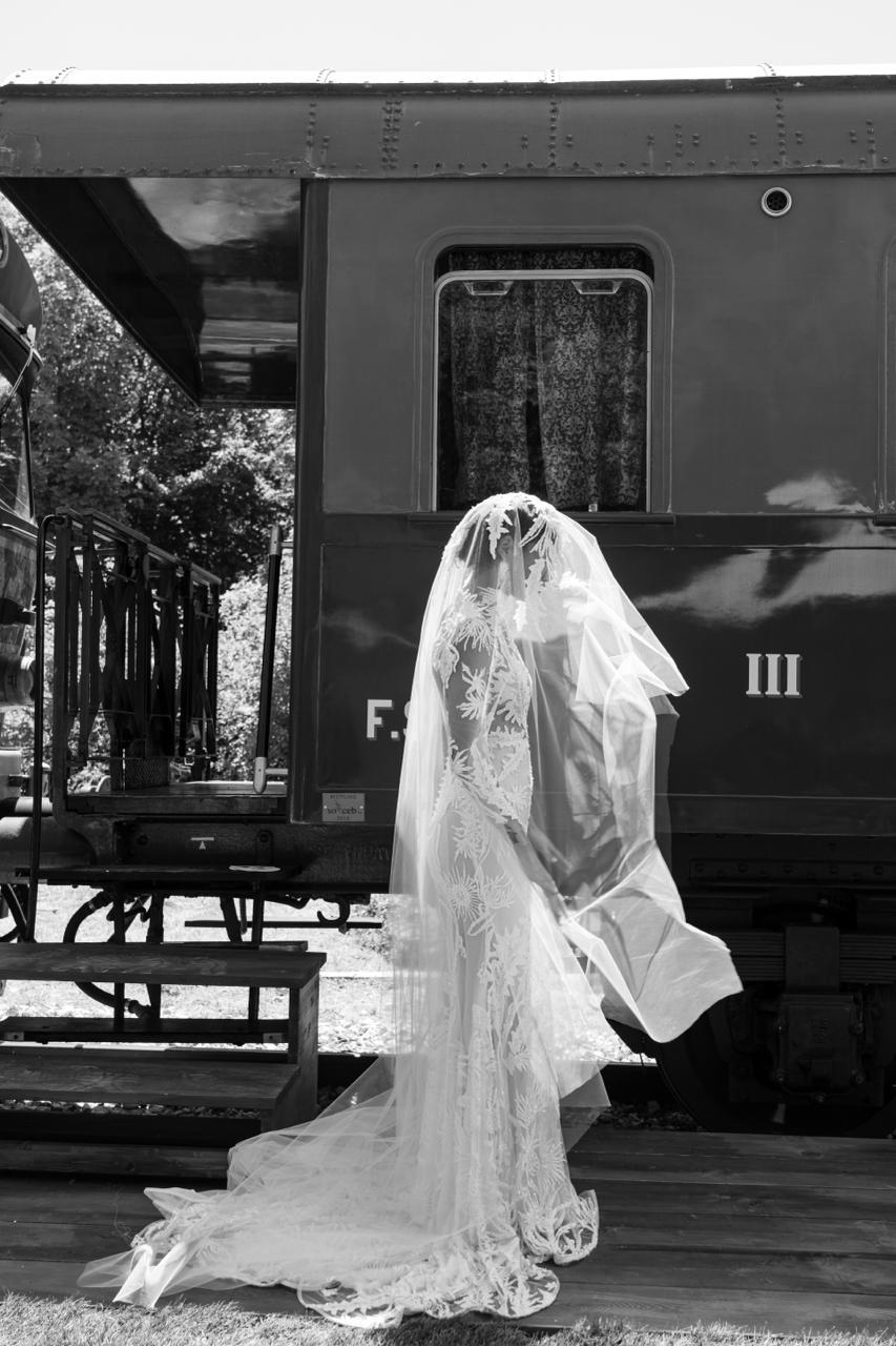 Sposa con lungo velo in abito Andrea Sedici accanto a una carrozza d’epoca, in bianco e nero