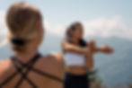 Women facing each other stretching their arm across their body with mountains in the background