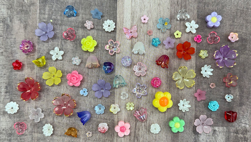 There is an array of flower beads spread out along a table. There are some that are opaque, translucent, shiny, matte, frosted. There are also a variety of sizes, colors, shapes, and kinds.  These include spring-blooming tulips and daffodils, summer-thriving hydrangeas and dahlias, and popular garden staples like roses, lilies, and daisies.
