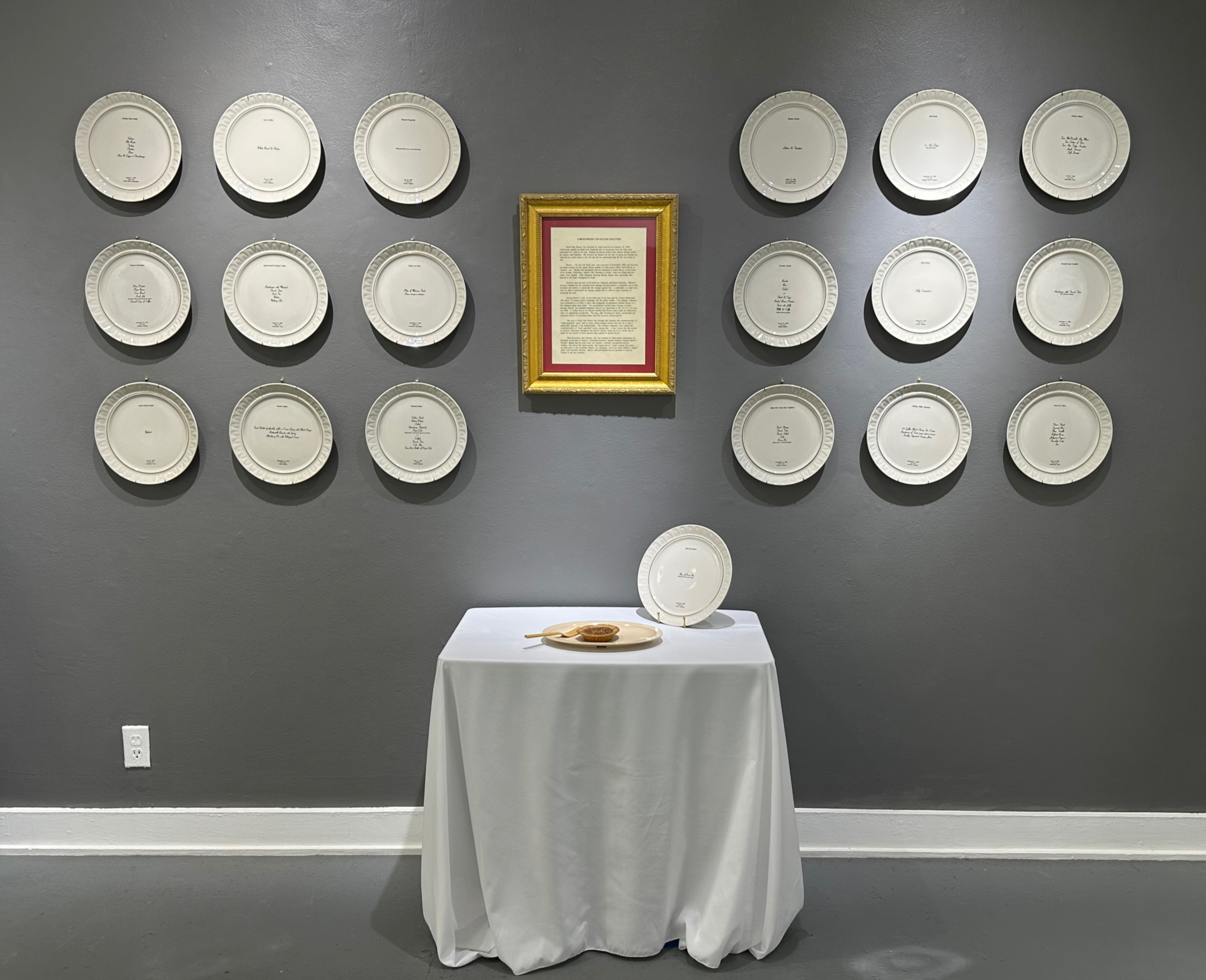 Mixed media installation which includes small table covered with white tablecloth.  One slice of pecan pie sits on a plastic dish.  A bone china dinner plate is standing. 18 bone china dinner plates hang on a gray wall.  Gold framed story about Ricky Ray Rector hangs centered on the wall behind the table.
