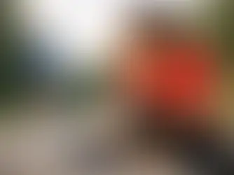 Woman in a red shirt sits on a path holding her knee in pain. A runner in blue is in the distance. Background is green and blurry.