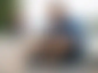 Man in blue shirt ties shoelaces beside crutches on a riverside path. A woman jogs in the background. Sunny, serene outdoor setting.