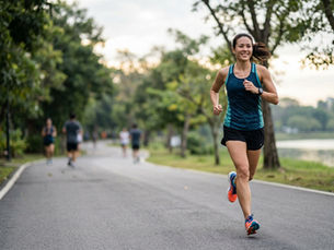 A woman in blue tank top runs on a tree-lined path, smiling. Others jog in the background by a lake, creating a lively, serene scene