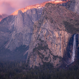 A photo of Bridalveil Fall at sunset in Yosemite National Park. The waterfall has already fallen into shadow while Half Dome is painted in sunlight through a break in low clouds.