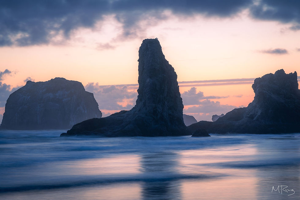 Seastacks rise from the ocean as soft waves reflect the colors of a serene sunset sky at Bandon Beach. By Michael Rung