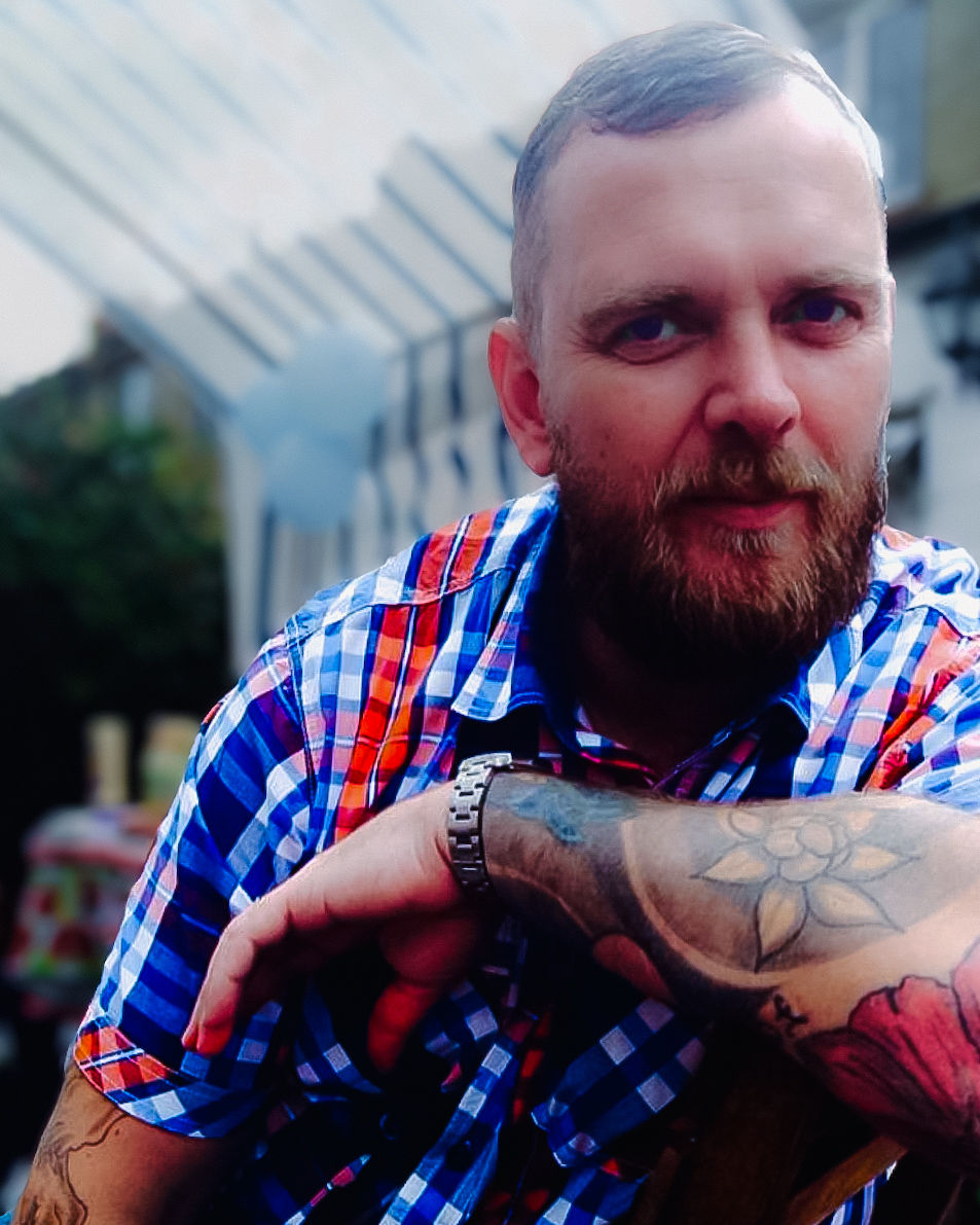 Tattooed Man in Checked Shirt – Relaxed Outdoor Portrait with Urban Backdrop