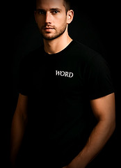 Young man in a dark room wearing a black shirt and jeans. The shirt has a single word on the left chest: word.