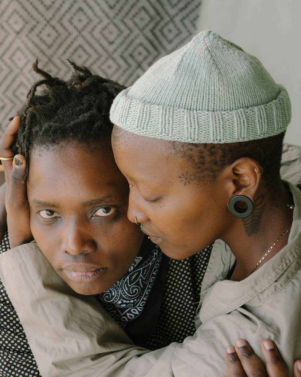 Two people embrace closely indoors, one in a pale knit beanie whispering to the other, with calm, intimate expressions.