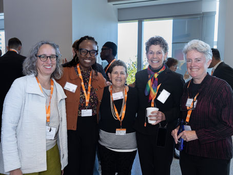 Five people smile at a conference, wearing orange lanyards with name tags. One holds a cup, another a bottle. Bright, modern interior.
