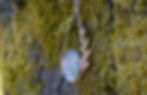 A large silver pendant hanging on o moss covered tree. The pendant is teardrop shape with a large oval stone set at the bottom. The stone is Flourite and a pale blue/purple colour. There is a simple frond of leaves going up the right side of the pendant.