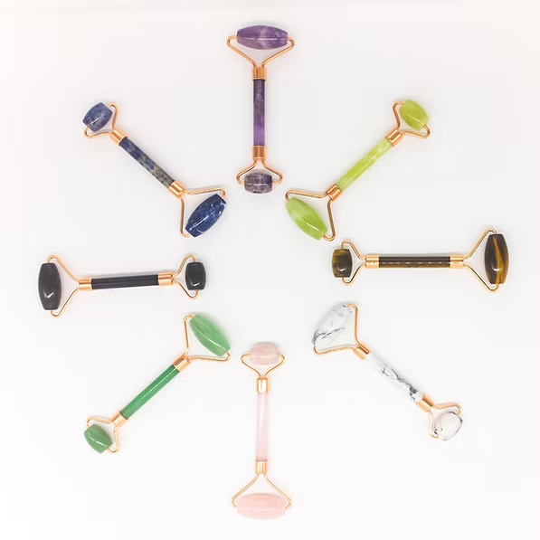 Seven colorful crystal facial rollers arranged in a circle on a white background, with purple, green, blue, black, white, and pink stones