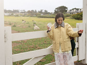 Woman in pale yellow jacket and white lace skirt standing by a white fence, holding a glass of white wine, with rolling green pasture and sheep grazing in the background under overcast coastal sky.