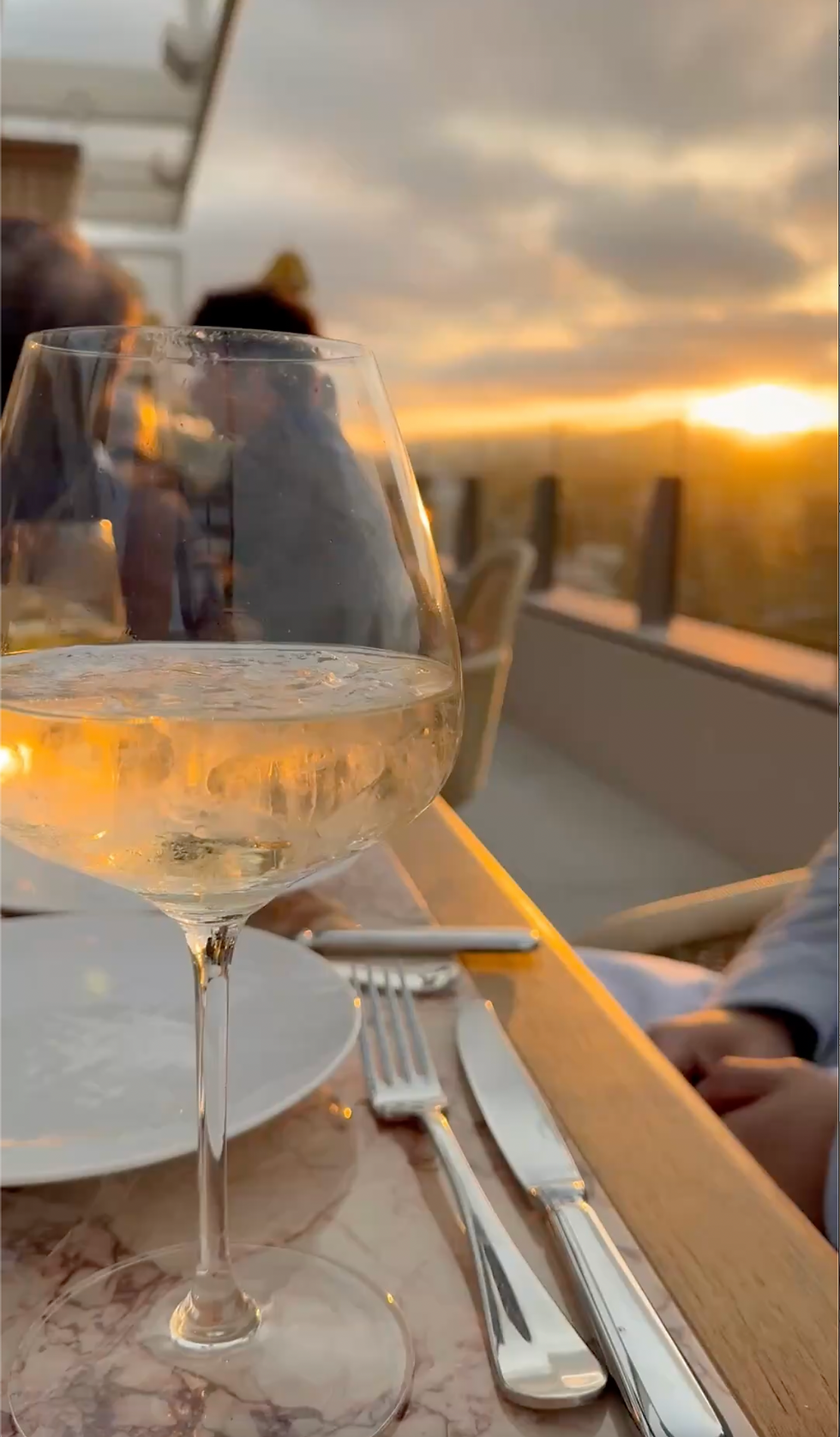 A glass of white wine catching the sunset on a rooftop dinner table, the sky glowing orange