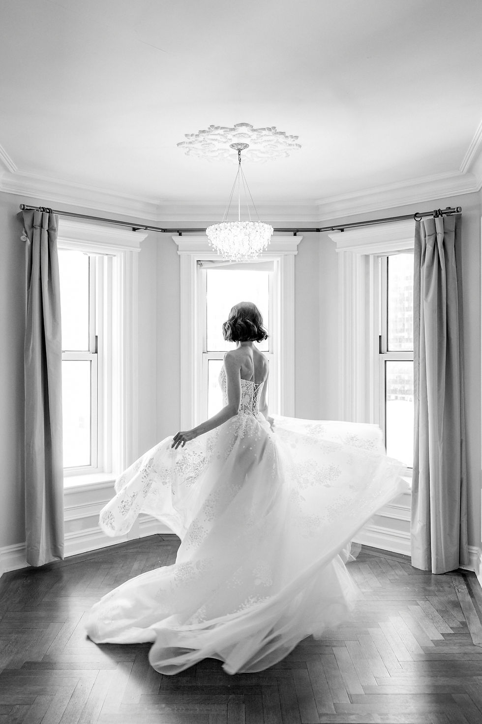 Bride in a flowing gown twirls in a bright room with large windows and elegant chandelier, creating a graceful and joyful atmosphere.