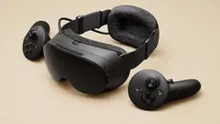 The Steam Frame VR headset in black, shown with its two matching controllers on a light beige surface. The headset has a soft fabric interior and an adjustable strap.