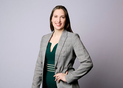 Juliette Dupré, a female business woman in a green dress and grey blazer smiling and looking at the viewer standing with left arm and hand on left hip.