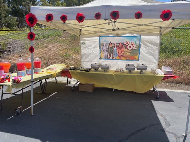 An outdoor tent setup decorated with large red poppies and a 'Wizard of Oz' banner. Tables underneath are covered with yellow tablecloths and filled with chafing dishes, drinks in dispensers, leis, and snacks, ready for a themed lunch event.