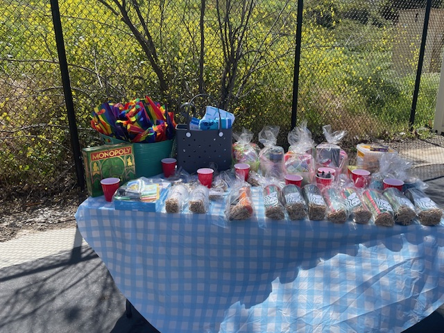 A table covered with a blue-and-white checkered cloth displaying a variety of raffle prizes including wrapped tumblers, Starbucks gift sets, red cups, a classic Monopoly game, rainbow umbrellas in a teal bin, and a dark blue tote bag. The setup is located outdoors near a fence with bright yellow wildflowers in the background.