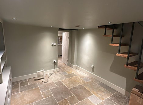 A recently painted room with a white ceiling and walls painted in a light grey colour. The room has a tiled floor with large, irregular tiles in shades of brown and beige. There are built-in shelves on the left side of the room, and a door leading to another room. A radiator is visible on the right side, and a set of stairs with wooden treads and metal railings leads up to the next level. The room is well-lit with recessed ceiling lights.