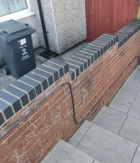 A new soldier course to brick wall with a black brick coping. Black cables run vertically down the wall. There are steps leading down to a paved area, which were recently installed.