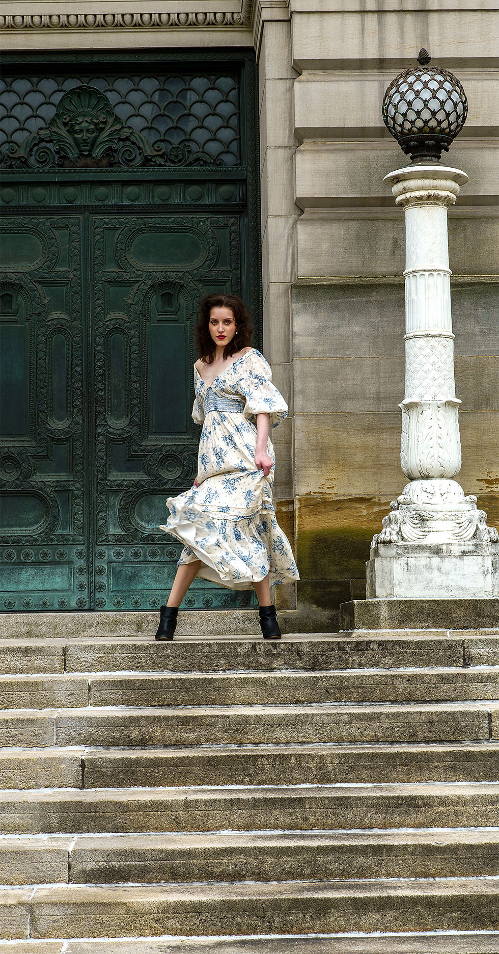 Vertical Spring Silhouette fashion image of a woman in a cream printed maxi dress with blue botanical motifs creating dress movement before classical stone architecture.