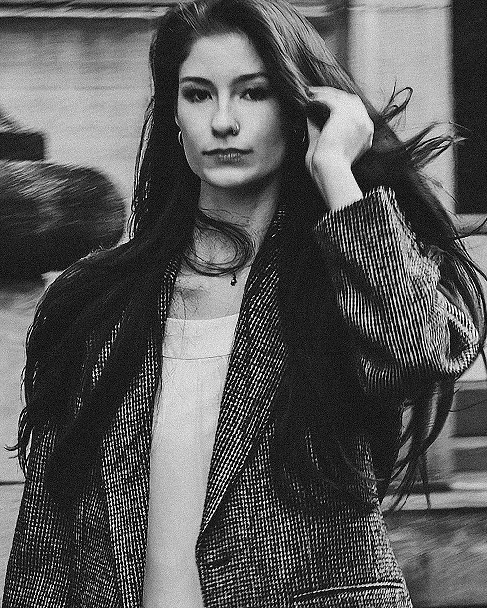 Black-and-white Autumn Structure fashion portrait of a woman in a tailored tweed blazer walking toward the camera and making eye contact on a city sidewalk.