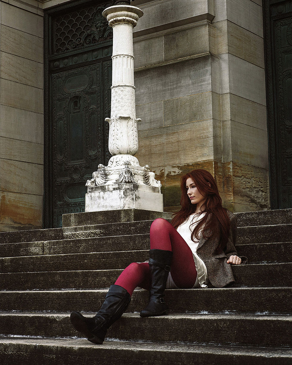 Reclining across stone steps, a woman in an tweed sport coat, wine-colored tights, and black boots is framed by a white column and green architectural doors.